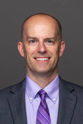 A man in a grey suit and purple shirt and tie smiles in to camera